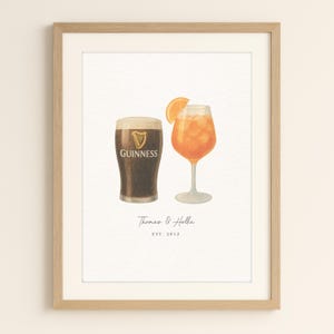 May include: A framed wall art print featuring a watercolour illustration of a dark Guinness stout glass next to an orange Aperol spritz cocktail with a slice of orange. Below the drinks, the text reads Thomas & Hollie and EST. 2012.