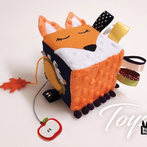Può includere: Un giocattolo per bambini a forma di cubo con un design a volpe, con tessuto di peluche arancione e bianco. Il giocattolo include nastri e un accessorio a forma di foglia e mela. La parola "Toys" è visibile. Il giocattolo è progettato con varie trame e colori.