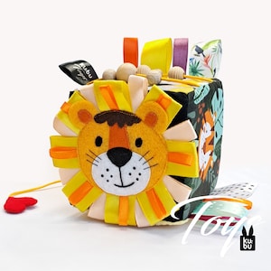 Può includere: Un giocattolo di stoffa a forma di cubo colorato con un viso di leone su un lato. Il cubo ha diverse texture e motivi, tra cui una scena della giungla e un motivo a pois bianco e nero. Il giocattolo è realizzato dal marchio "Kubu Toys".