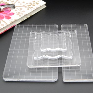 May include: Three clear acrylic stamping blocks with a grid pattern. The largest block is on the bottom, followed by a smaller block, and then the smallest block on top. The blocks are designed for use with clear stamps.
