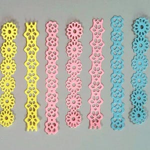 May include: Eight strips of decorative paper lace in yellow, pink and blue. The lace is in a delicate floral pattern.