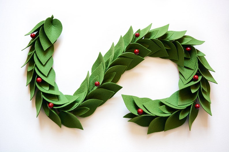 May include: A green felt garland with red felt berries. The garland is made of individual felt leaves that are sewn together. The garland is in the shape of a circle.