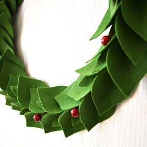 May include: A green felt wreath with red berries. The wreath is made of individual felt leaves that are layered to create a full and lush look.