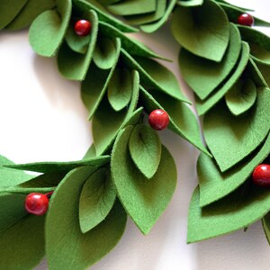 May include: A green felt wreath with red felt berries. The wreath is made of overlapping leaves and is perfect for a holiday or winter decor.