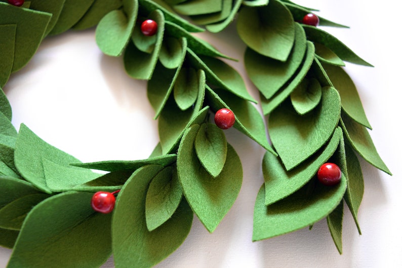 May include: A green felt wreath with red felt berries. The wreath is made of overlapping leaves and is perfect for a holiday or winter decor.