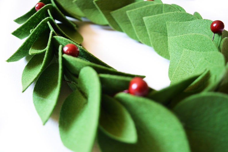 May include: A green felt wreath with red felt berries. The wreath is made of individual felt leaves that are sewn together.