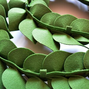 May include: A close-up of a green felt garland made of individual leaves. The leaves are sewn together to create a continuous garland.
