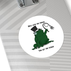 May include: A round sticker featuring a cartoon frog in a top hat and glasses, holding a whip. The frog is green, and the sticker has the text "MESS WITH THE FROGGO, YOU GET THE FLOGGO".