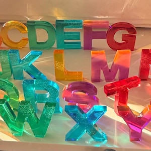 May include: A complete set of twenty-six individual alphabet letters made from translucent, multi-colored resin. Each letter features a glossy finish and vibrant hues including red, orange, yellow, green, blue, purple, and pink, arranged in order.