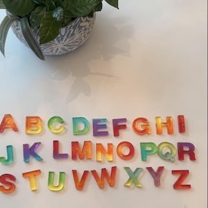 May include: A complete set of twenty-six alphabet letters made from translucent, multi-colored resin. Each letter features a smooth, glossy finish with blended gradients of red, orange, yellow, green, blue, and purple hues arranged in three rows.