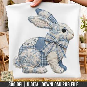 May include: A white pillow featuring a patchwork bunny design. The bunny is primarily blue and white, with floral and gingham patterns. It has a large bow and lace accents, and a black button eye. The pillow is held up by a hand.