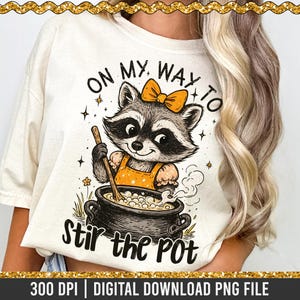 Puede incluir: Camiseta color crema con un mapache de dibujos animados con un lazo revolviendo una olla. El texto "ON MY WAY TO Stir the POT" está encima y debajo de la ilustración del mapache. El diseño incluye estrellas y flores.
