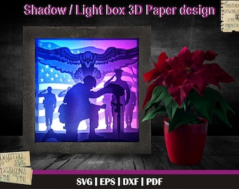 May include: A 3D papercraft design of a soldier kneeling in front of an American flag with an eagle flying above. The design is illuminated with blue and purple lights. The design is in a shadow box frame.