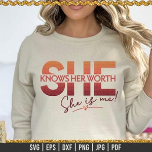 Può includere: Una felpa beige con un design grafico che dice "SHE KNOWS HER WORTH She is me!" in un gradiente rosso, arancione e giallo. Il testo è in grassetto, carattere sans-serif.