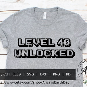May include: A gray t-shirt with the text "LEVEL 40 UNLOCKED" in a pixelated font. The text is black and white and is centered on the shirt. The shirt is on a wooden background.