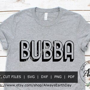 Puede incluir: Una camiseta gris con la palabra "BUBBA" impresa en negro, en letras en 3D. La camiseta está sobre un fondo de madera blanca. El texto "ALWAYS EARTH DAY AED" está en un círculo con hojas verdes.