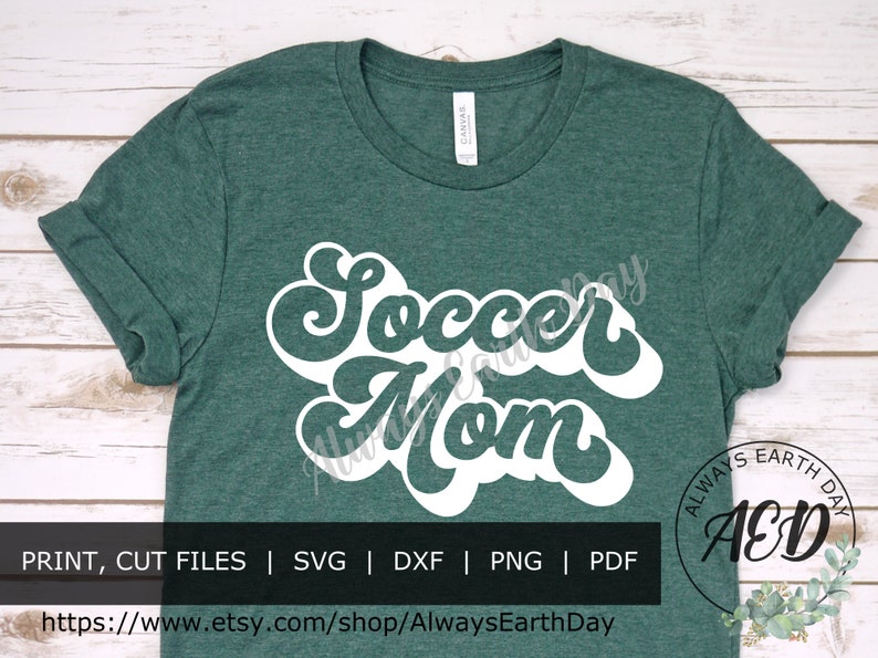 May include: A green t-shirt with white text that reads "Soccer Mom" in a retro font. The text is in a large, bold font and is centered on the shirt. The shirt is on a white wooden background. The text "ALWAYS EARTH DAY AED" is in a circle on the bottom right corner of the image.