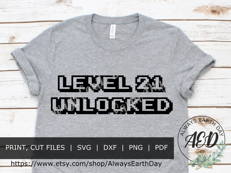 May include: A gray t-shirt with the text "LEVEL 21 UNLOCKED" in a pixelated font. The text is black and the shirt is a heather gray color. The text is centered on the shirt. The shirt is a standard t-shirt style with short sleeves and a crew neck.
