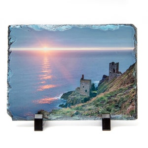 May include: A slate coaster featuring a photograph of a sunset over the sea with two old stone buildings on a cliffside.