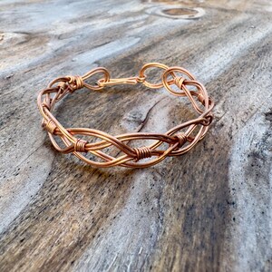 Op de afbeelding: Een koperkleurige draadarmband met een geweven ontwerp. De armband heeft een reeks in elkaar grijpende draden, waardoor een textuurpatroon ontstaat. De sluiting bestaat uit twee verbonden lussen. De armband wordt gepresenteerd op een verweerd houten oppervlak.