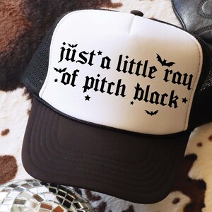 Puede incluir: Una gorra de camionero blanca y negra con una parte trasera de malla negra. La parte delantera de la gorra tiene el texto "just a little ray of pitch black" en una fuente g&oacute;tica con murci&eacute;lagos y estrellas.