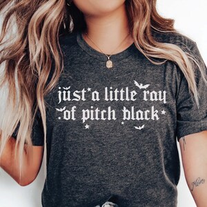 Puede incluir: Una camiseta gris oscuro con el texto "just a little ray of pitch black" en blanco en fuente g&oacute;tica. El texto est&aacute; rodeado de peque&ntilde;as estrellas y murci&eacute;lagos.