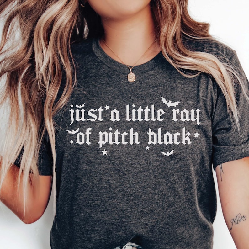 Puede incluir: Una camiseta gris oscuro con el texto "just a little ray of pitch black" en blanco en fuente g&oacute;tica. El texto est&aacute; rodeado de peque&ntilde;as estrellas y murci&eacute;lagos.
