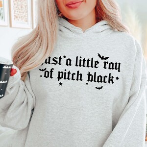 Puede incluir: Sudadera gris con capucha con el texto "Just a little ray of pitch black" en fuente g&oacute;tica negra. El texto est&aacute; rodeado de peque&ntilde;as estrellas y murci&eacute;lagos.