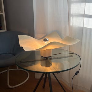 May include: A table lamp featuring a wide, wavy, off-white shade with a textured surface. The lamp has a central silver-capped light bulb and a square white base. A black power cord with an inline switch extends from the base of the lamp.
