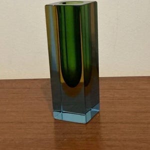 Puede incluir: Un jarrón de vidrio alto y rectangular con una superficie lisa y pulida. El vidrio muestra una transición de color degradada, que cambia de un verde profundo y vibrante en la parte superior a un tono azul claro transparente en la base.