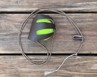May include: A brown leather tennis ball holder with a green tennis ball inside. The holder is attached to a brown cord with a white string attached to the end.