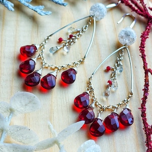 以下が含まれることがあります： シルバーカラーのドロップピアス。各イヤリングは、ファセット加工された濃い赤色の宝石のクラスターを備えた涙の形をしたフレームを備えています。小さな透明と赤のビーズがチェーンからぶら下がり、大きな質感のある透明なビーズが各イヤリングを飾っています。