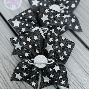 May include: Black and silver glitter hair bows with a star pattern and a planet embellishment. The bows are arranged on a wooden surface. The bows have a glittery texture and are likely for a child.