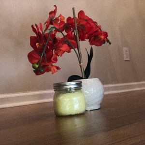 May include: A clear glass jar with a yellow lid filled with a yellow substance. The jar is sitting on a wooden floor next to a white ceramic planter with a red orchid plant.