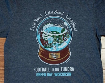 Könnte beinhalten: Ein dunkelblau meliertes Kurzarm-T-Shirt mit einer Schneekugel-Grafik, die ein Football-Stadion, Tannenbäume und einen Schneemann zeigt. Der Text lautet Let it Snow! Let it Snow! Let it Snow! FOOTBALL IN THE TUNDRA GREEN BAY, WISCONSIN.