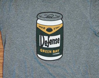 Könnte beinhalten: Graues T-Shirt mit einem Aufdruck einer Dose Green Bay Packers "Defense" Bier. Die Dose ist grün und gelb mit den Worten "Open Up A Can!" und "Green Bay It's Strong" bedruckt.