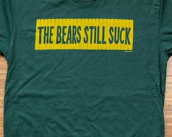 Könnte beinhalten: Ein dunkelgrünes T-Shirt mit dem Text "THE BEARS STILL SUCK" in Gelb auf der Vorderseite. Das Shirt hat ein kleines Etikett auf der Vorderseite, auf dem "Tundra Town" steht.
