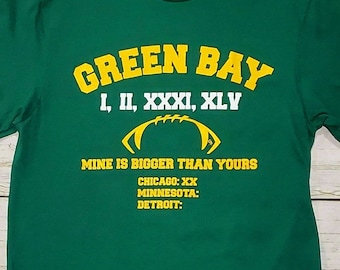 Könnte beinhalten: Grünes T-Shirt mit einer gelben Grafik, auf der "Green Bay I, II, XXXI, XLY" und "Mine is bigger than yours" mit einem Football-Motiv steht. Der Text "Chicago: XX, Minnesota: Detroit:" befindet sich ebenfalls auf dem Shirt.