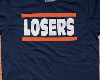 Könnte beinhalten: Ein marineblaues T-Shirt mit dem Wort "LOSERS" in weißen Buchstaben, über und unter dem Wort befinden sich zwei orangefarbene Streifen.