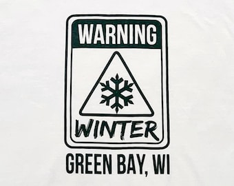Könnte beinhalten: Weißes T-Shirt mit einem schwarzen und grünen Warnschild mit der Aufschrift "WARNING WINTER GREEN BAY, WI". Das Schild hat ein Schneeflocke in einem Dreieck.