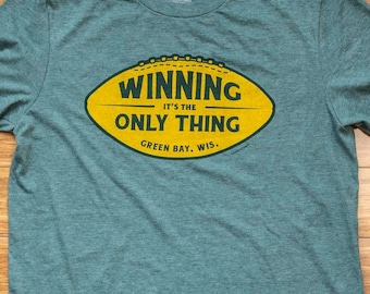 Könnte beinhalten: Ein blaugrünes T-Shirt mit einem gelben Football-Motiv, auf dem "Winning, It's the Only Thing, Green Bay, Wis." steht.