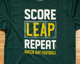 Könnte beinhalten: Grünes T-Shirt mit einer weißen Grafik, auf der "SCORE LEAP REPEAT GREEN BAY FOOTBALL" im Stil einer alten Anzeigetafel steht.
