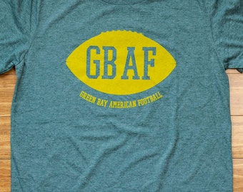 Könnte beinhalten: Ein tealgrünes T-Shirt mit einem gelben Football-Motiv, auf dem "GB AF" und darunter "Green Bay American Football" steht.
