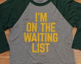 Könnte beinhalten: Ein grau-grünes Baseball-T-Shirt mit dem Aufdruck "I'm on the waiting list" in Gelb auf der Vorderseite.
