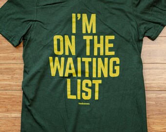Könnte beinhalten: Ein dunkelgrünes T-Shirt mit dem Text "I'm on the waiting list" in gelber Farbe. Der Text "tundratown" ist in kleineren gelben Buchstaben unter dem Haupttext gedruckt.