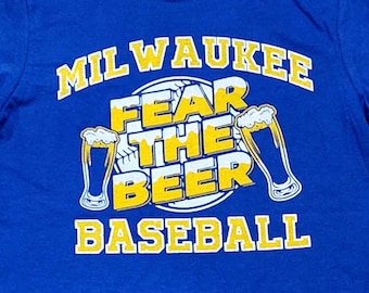 Könnte beinhalten: Ein königsblaues T-Shirt mit dem Schriftzug "MILWAUKEE BASEBALL" in weißer und gelber Schrift. Der Satz "FEAR THE BEER" ist zentriert, mit Bierkrügen auf beiden Seiten. Das T-Shirt hat einen Rundhalsausschnitt und ein kleines "S"-Größenetikett.