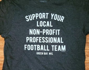 Könnte beinhalten: Ein dunkelgraues T-Shirt mit weißem Text, der "Support Your Local Non-Profit Professional Football Team Green Bay, Wis." lautet.