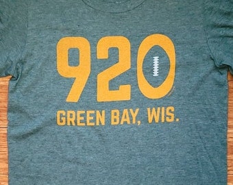 Könnte beinhalten: Grünes Kurzarm-T-Shirt mit dem Text "920 GREEN BAY, WIS." in Gelb. Die Zahl "920" befindet sich über einer weißen Football-Grafik. Das Shirt hat ein kleines "S"-Größenetikett und das "Tundra Town"-Logo.