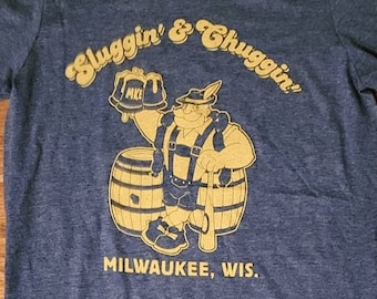 Könnte beinhalten: Ein blaues T-Shirt mit einer gelben Grafik eines Cartoon-Mannes, der ein Bierkrug und einen Baseballschläger hält. Der Text "Sluggin' & Chuggin'" befindet sich über dem Cartoon und "Milwaukee, Wis." unter dem Cartoon.