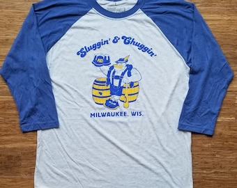 Könnte beinhalten: Ein weißes und blaues Baseball-T-Shirt mit einer Cartoon-Illustration eines Mannes, der zwei Biere hält und neben zwei Fässern steht. Der Text "Sluggin' & Chuggin'" befindet sich über der Illustration und "MILWAUKEE, WIS." darunter.